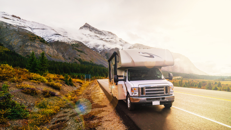 A white RV travels along a scenic mountain road with snow-capped peaks, green trees, and golden foliage under a bright, sunny sky.