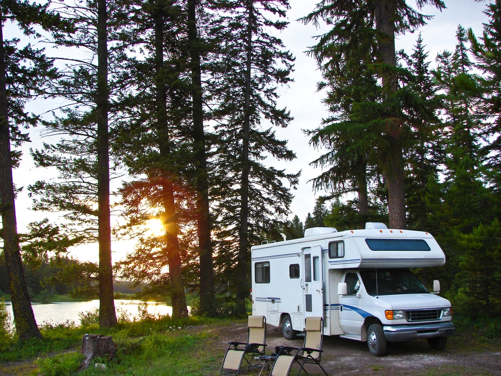 A white RV is parked among tall pine trees near a lake at sunset, with two folding chairs set up outside on a grassy clearing.